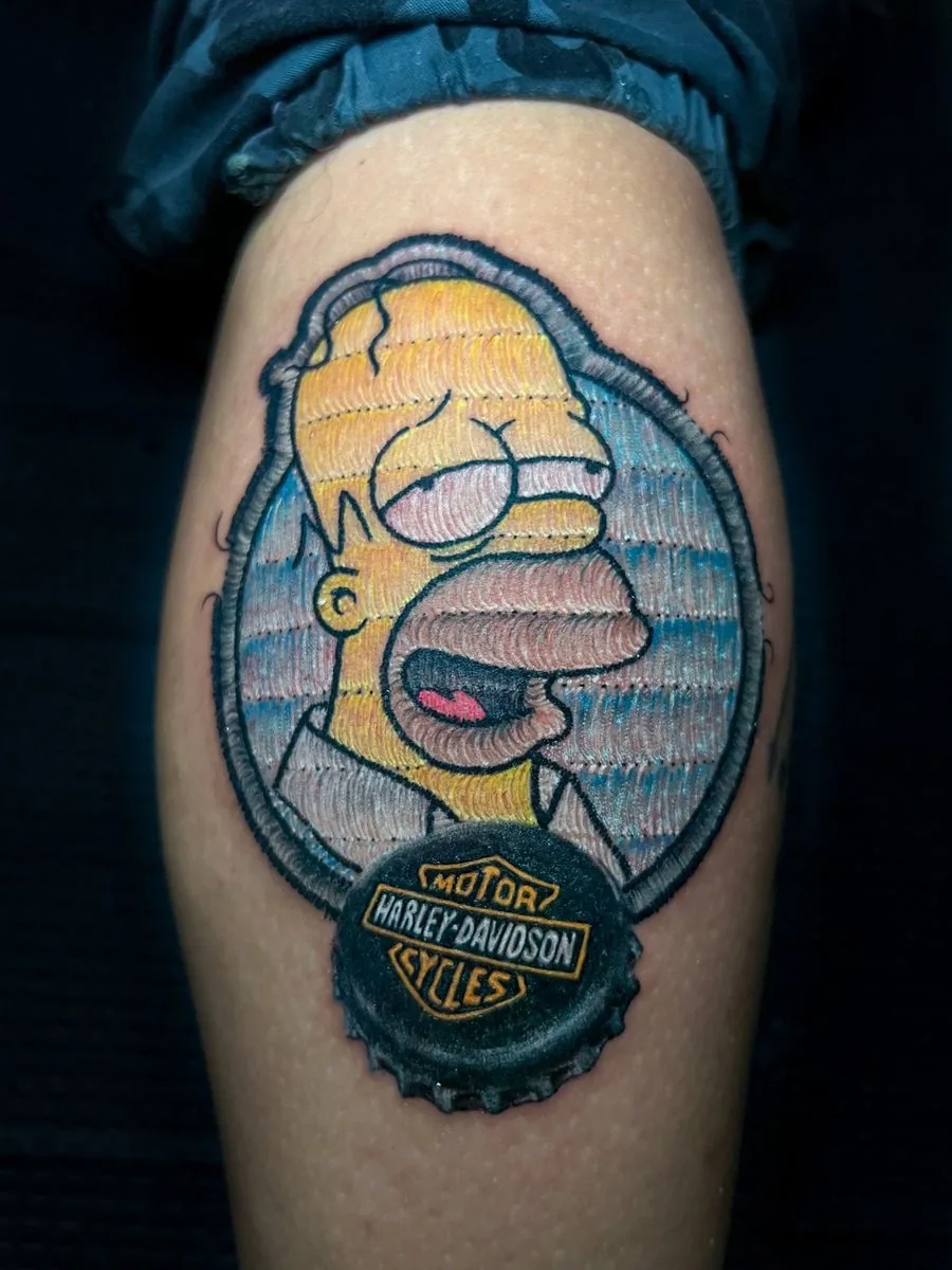Tatuaje estilo parche bordado: personaje de caricatura con borde bordado y una chapa de botella, en la pierna · Wilson Escobar, tatuador en Miami
