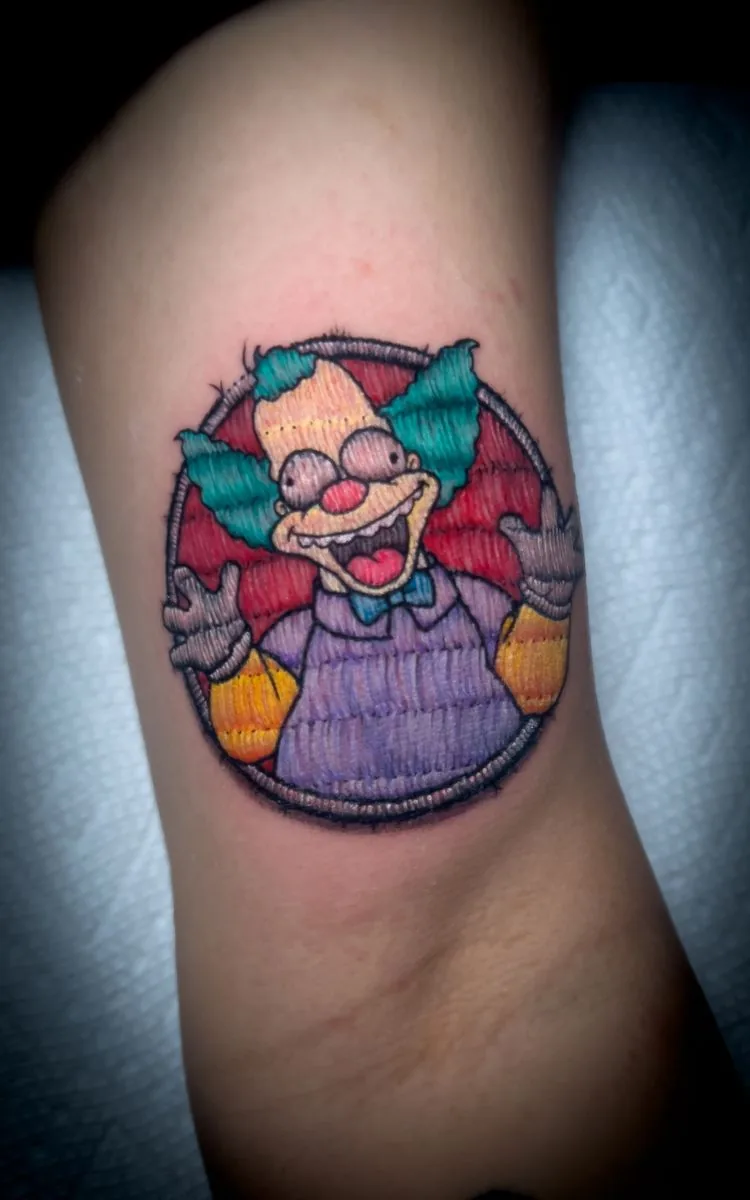 Tatuaje estilo parche bordado: payaso de caricatura en un parche redondo, en el brazo · Wilson Escobar, tatuador en Miami
