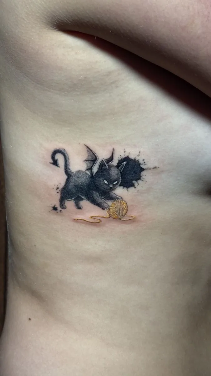 Tatuaje ilustrativo: gatito negro con alas de murciélago jugando con un ovillo de lana, en las costillas · Wilson Escobar, tatuador en Miami